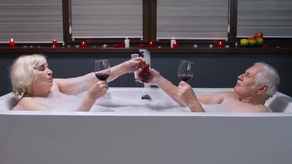 Senior Couple Man and Woman Lying in Warm Bath with Bubbles, Enjoying Relaxation, Drinking Red Wine
