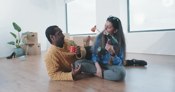 Young couple in new home sitting on the floor holding plant and drill