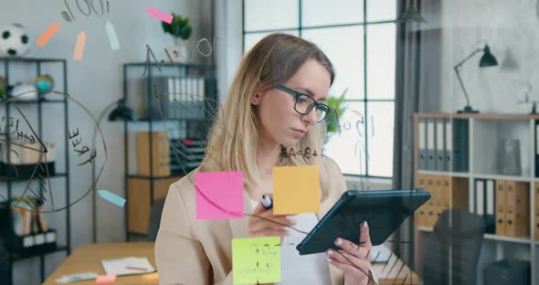 Woman in Glasses which Standing Near Glass Board and Writing Datas from Tablet PC