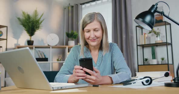 Senior Woman Using Mobile While Sitting at Desk with Laptop