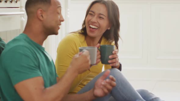 Happy biracial couple sitting on kitchen's floor, drinking coffee and talking