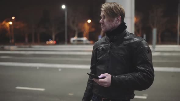 A Young Man Wearing a Leather Jacket Sits on His Motorcycle at the Sunset and Speaks on the Phone at