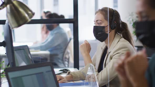 Caucasian businesswoman wearing face mask at desk with sneeze shield talking wearing phone headset