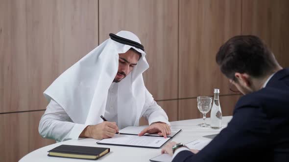 Arab Man in a White Kandura Sign Financial Documents with a Partner Businessman in a Suit While