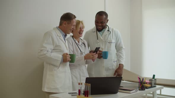 Positive Diverse Multiracial Medical Staff Relaxing After Work and Sharing Phone at Medical Office