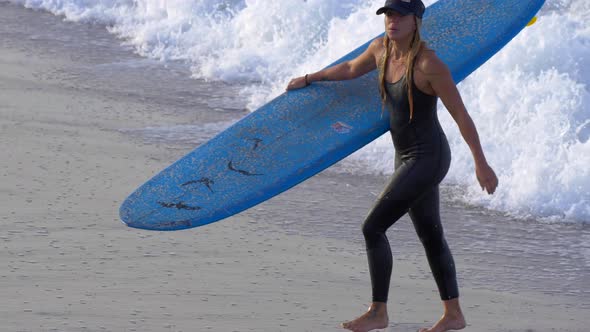 A young woman in a wetsuit walking with her longboard surfboard.