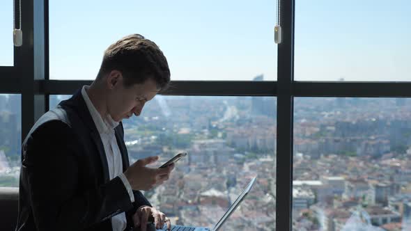 Businessman Is Working with Phone and Laptop Sitting Near the Window with City View.