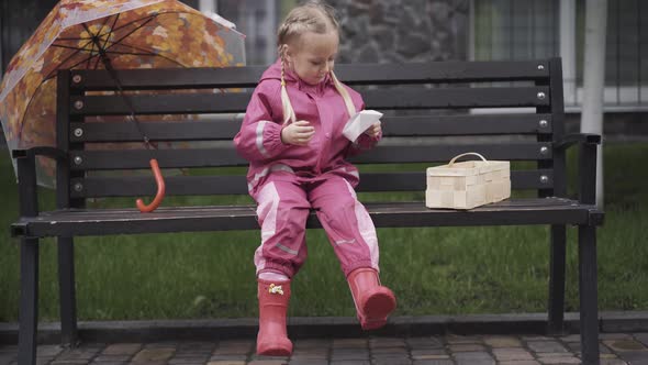 Joyful Caucasian Girl Sitting on Bench and Playing with Paper Boat. Wide Shot of Happy Little Child