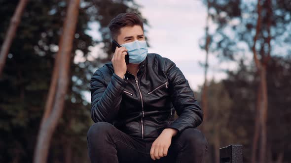 Portrait of Young Caucasian Man in Medical Face Mask Talking on the Phone in City Park at Sunny Day.