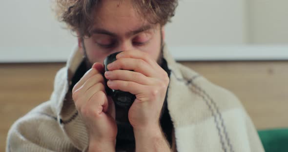 Ill Man Blowing on Cup with Hot Tea and Drinking While Sitting on Couch at Home