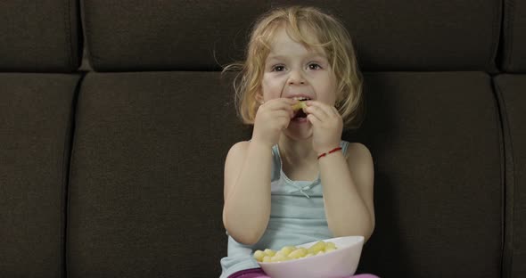 Girl Sitting on Sofa and Eating Corn Puffs. Child Watching Tv, Taste Puffcorns