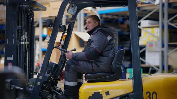 Wide Shot Side View of Male Warehouse Loader Sitting in Forklift Talking Looking Around