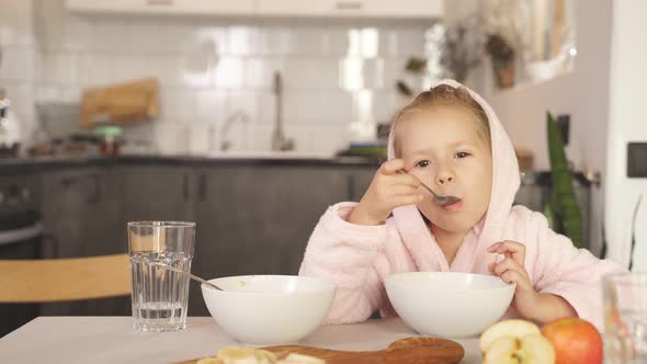 Girl with Blonde Hair Dressed in a Robe and Hood After a Shower Sits at the Table and Eats Oatmeal