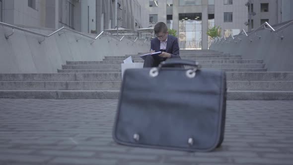 Handsome Well-dressed Boy Sitting on the Stairs on the Street Doing Home Work at the Background