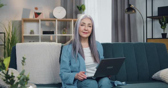 Senior Woman which Sitting on the Couch with Laptop and Poses on Camera