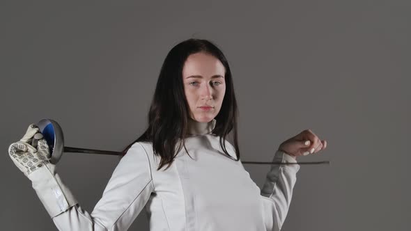 Portrait of a Young Woman Fencer in a White Uniform Posing with a Rapier in Her Hands on a Gray