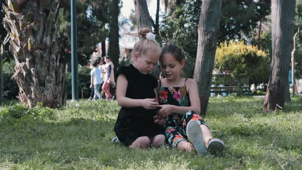 Two Little Girls Sitting on the Grass and Playing with the Phone