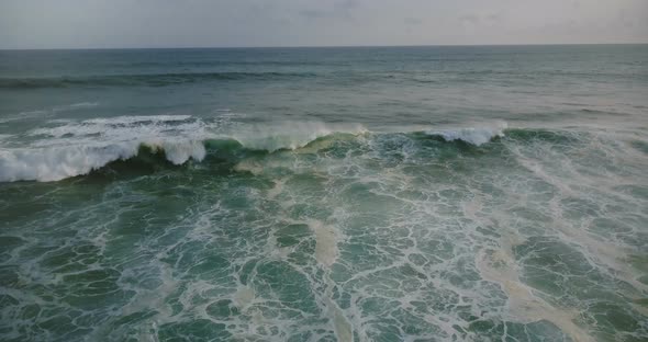 Drone Tilts Down To Follow Big Blue Ocean Wave Reaching the Shore and Crashing with White Seafoam