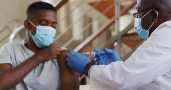 African american senior male doctor giving covid vaccine to male patient in home, wearing face masks