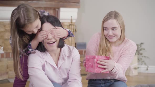 Two Caucasian Daughters Giving Surprise Present To Their Adorable Mother. Happy Brunette Woman