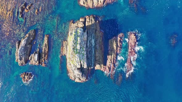 Aerial vertical high angle view of big rocks in sea coast while waves break against them at sunset