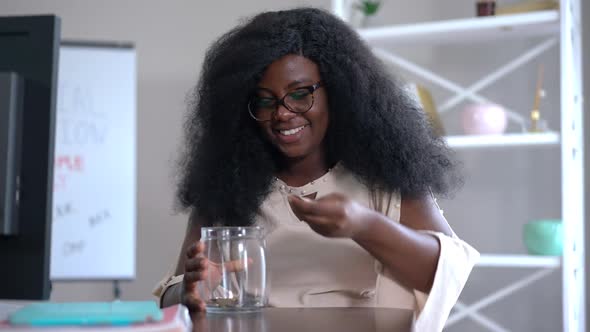 Joyful African American Female Numismatist Smiling Putting Coins in Glass Jar in Slow Motion Sitting