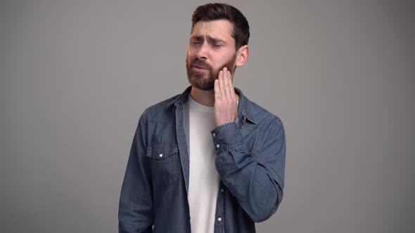 Frustrated bearded caucasian man touching cheek and wincing in pain feeling terrible tooth ache