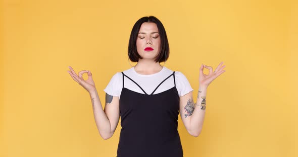 Young hipster woman with tattoos meditates and relaxes on a yellow background.