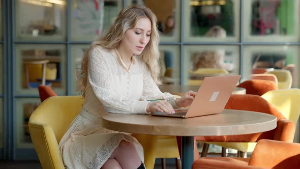Concentrated Young Caucasian Woman Typing on Laptop Keyboard and Drinking Coffee in Restaurant