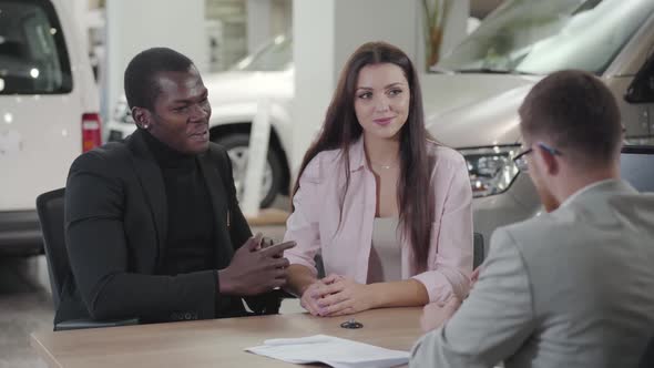 Happy Young Multiracial Couple Sitting in Car Dealership and Talking with Salesman