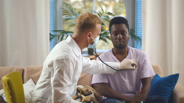 Young Doctor in Safety Mask Checking Heartbeat of Patient Using Stethoscope at Home