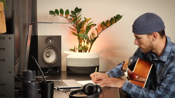 Young musician composer with acoustic guitar composing music or song, making notes in notebook.