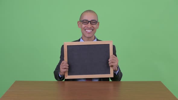 Happy Bald Multi Ethnic Businessman Talking While Holding Clipboard Behind Desk