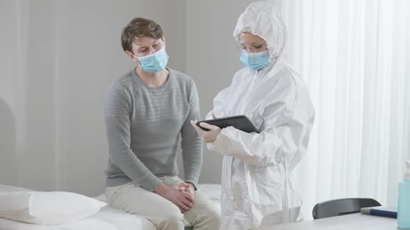 Professional Concentrated Woman in Protective Suit Using Tablet Consulting Ill Man Sitting on