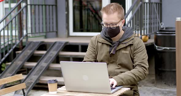 A man in a mask works on a laptop while sitting in a cafe on the street