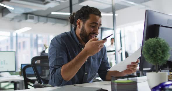 Mixed race businessman sitting at desk holding document, using smart phone and smiling