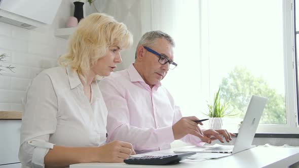 Senior Wife and Elderly Husband Counting Home Finance in Apartment Kitchen Next To Window