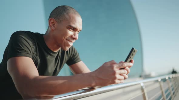 Cheerful bald sportsman wearing black t-shirt texting on mobile outdoors