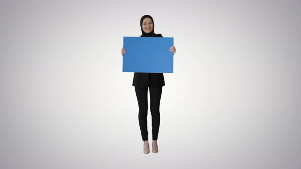 Smiling Arab Woman in Hijab Holding Blank Blue Poster and Looking at It on Gradient Background