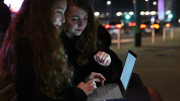 two young women outdoor city night using computer laptop