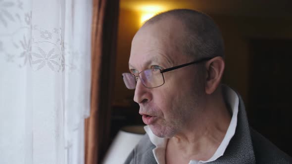 An Elderly Man at the Window Takes a Pill and Drinks It with Water