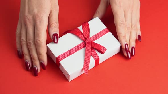 Woman Hands Take White Gift Box with Red Bow on Red Table Decorated with Heart