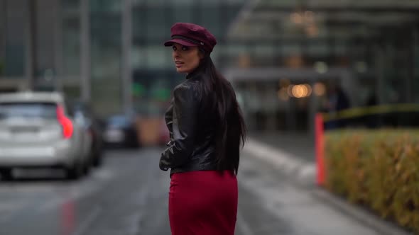 a Brunette with Long Hair in a Black Leather Jacket a Red Skirt and a Burgundy Cap Walks Against the