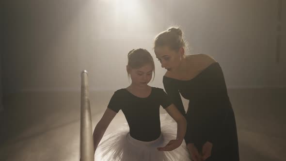 Medium Shot of Slim Woman Explaining Third Ballet Position Talking with Girl in Dance Studio Backlit