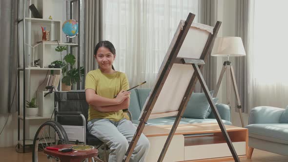 Asian Artist Girl In Wheelchair Holding Paintbrush And Cross His Arms While Painting On The Canvas