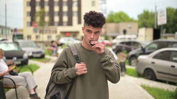 A young man with a backpack walks along the city alley and smokes