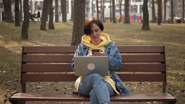 Young woman sitting on bench in spring park using laptop and smartphone