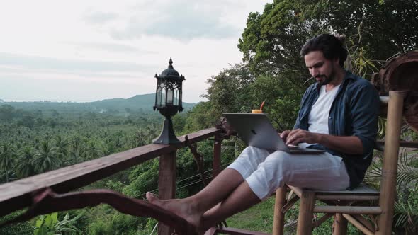 A young, handsome man with a beard works on a laptop in a cafe overlooking an incredible sunset.