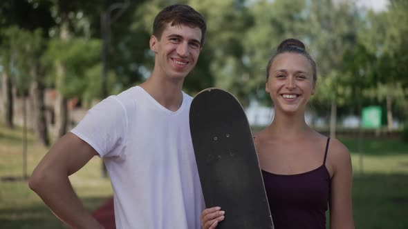 Happy Loving Young Couple with Skateboard Looking at Each Other Turning to Camera Gesturing Thumbs