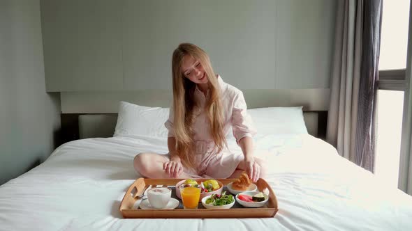 Happy Smiling Woman Eating Croissant on Breakfast in Bed in Modern Hotel